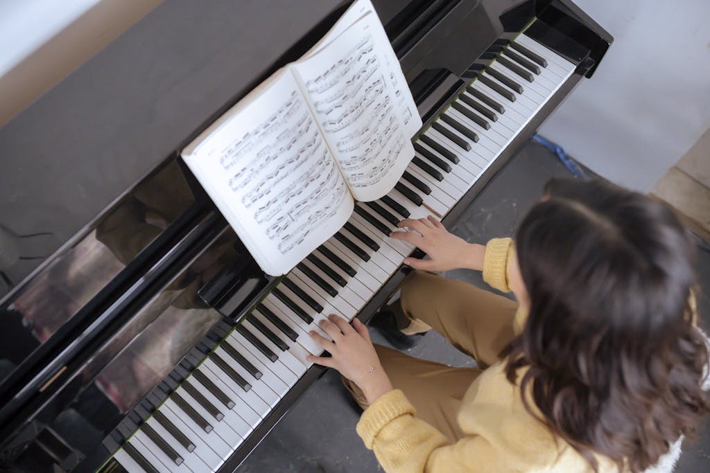 From above of crop faceless woman in warm yellow sweater with music book practicing music on piano in light classroom